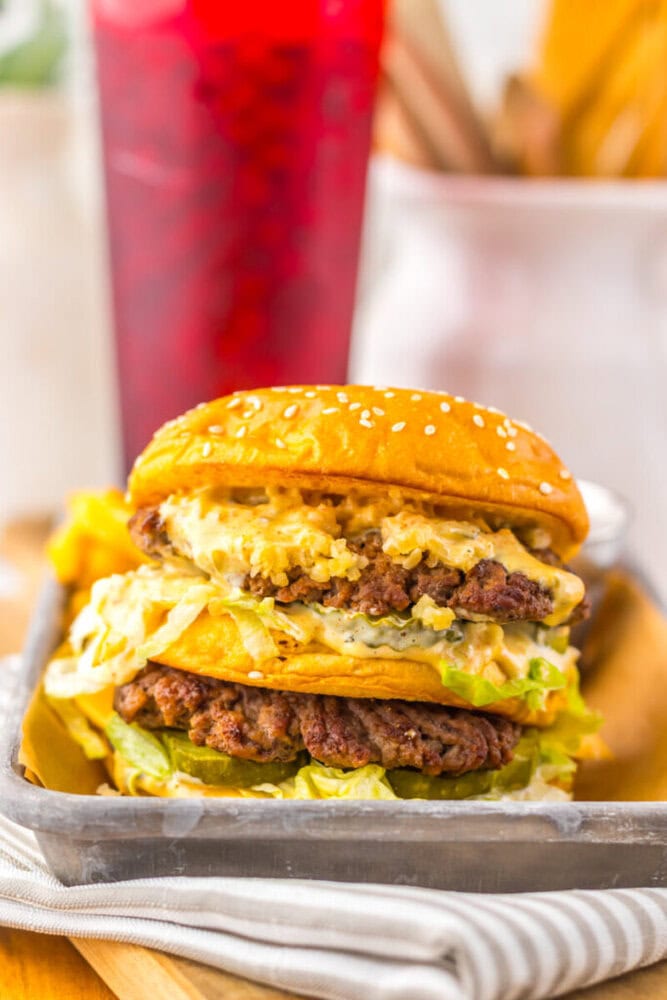 A double cheeseburger with lettuce, pickles, and sauce on a sesame seed bun sits on a metal tray, with a red drink in the background.