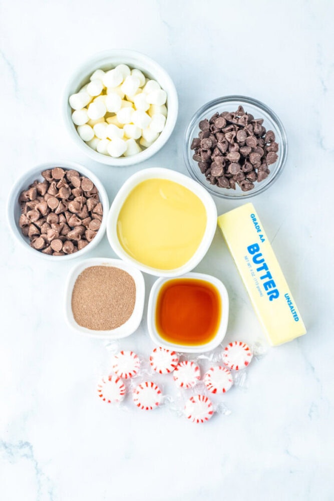 Bowls of white chocolate chips, milk chocolate chips, and chocolate chips, sweetened condensed milk, butter, vanilla extract, hot cocoa mix, and peppermint candies on a white surface.