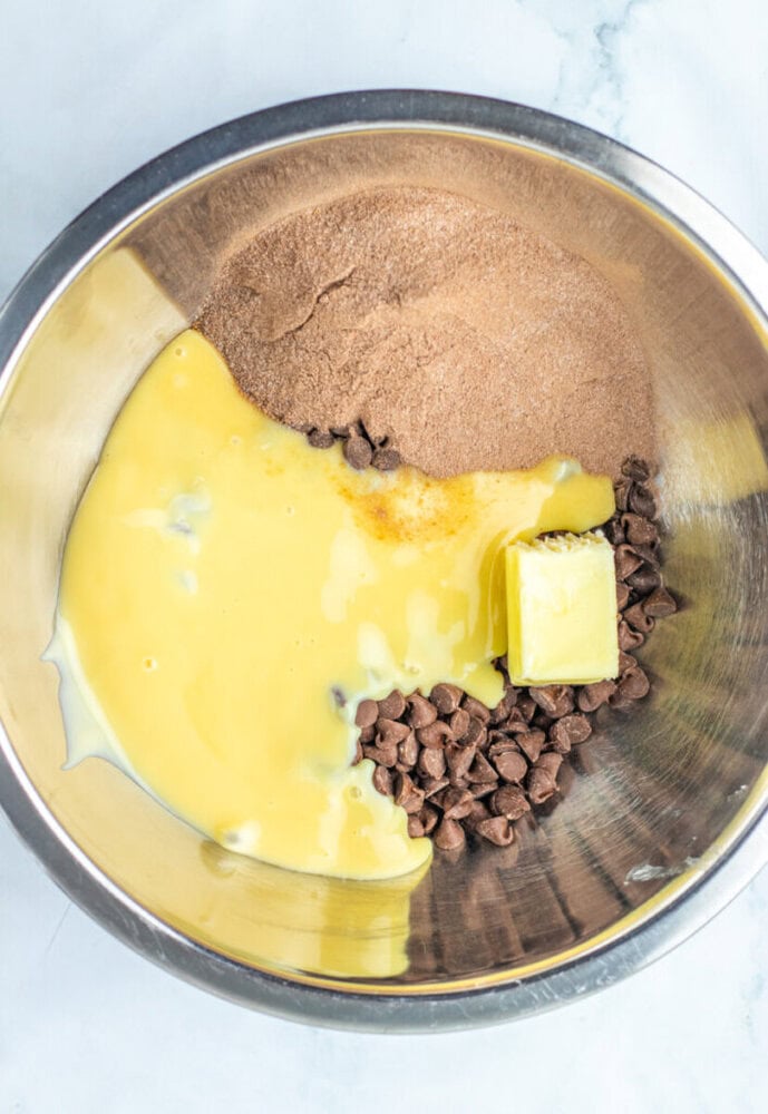 A metal bowl containing sweetened condensed milk, butter, chocolate chips, and brown sugar on a white surface.