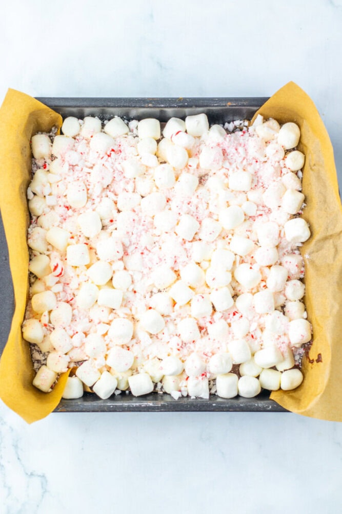 A square baking pan lined with parchment paper is filled with mini marshmallows and crushed peppermint pieces on top of a dessert base.