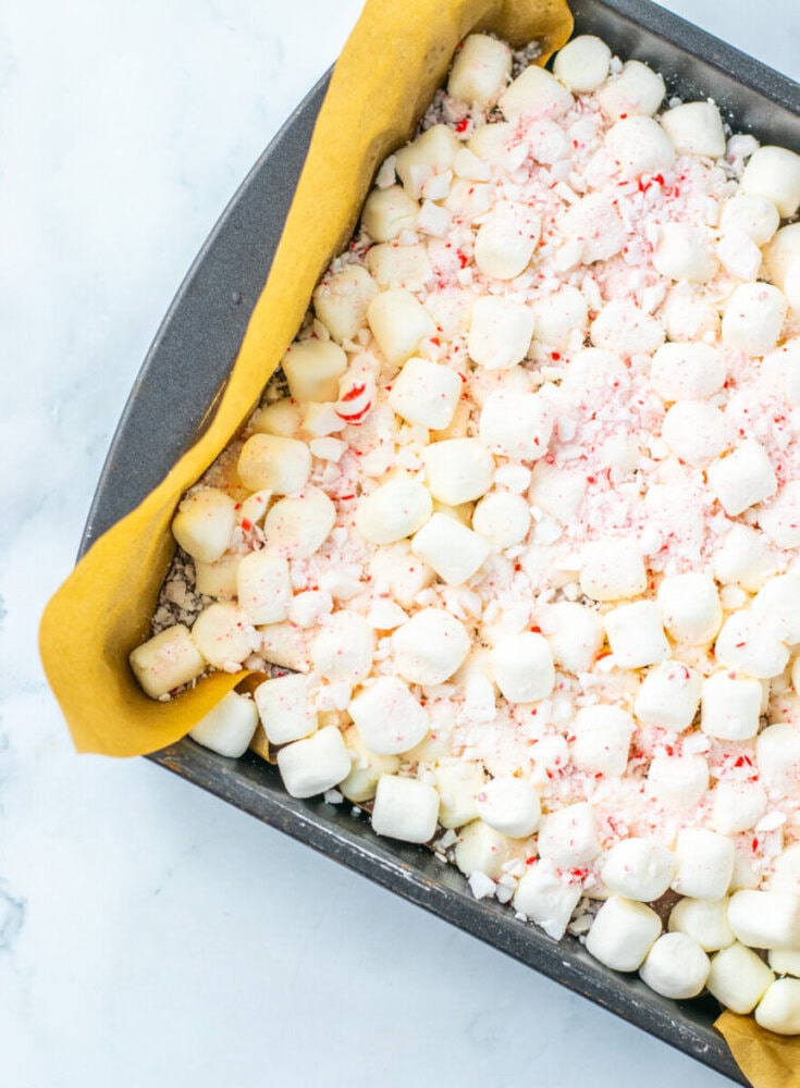 A baking pan filled with mini marshmallows and crushed peppermint on top of a yellow parchment paper lining.
