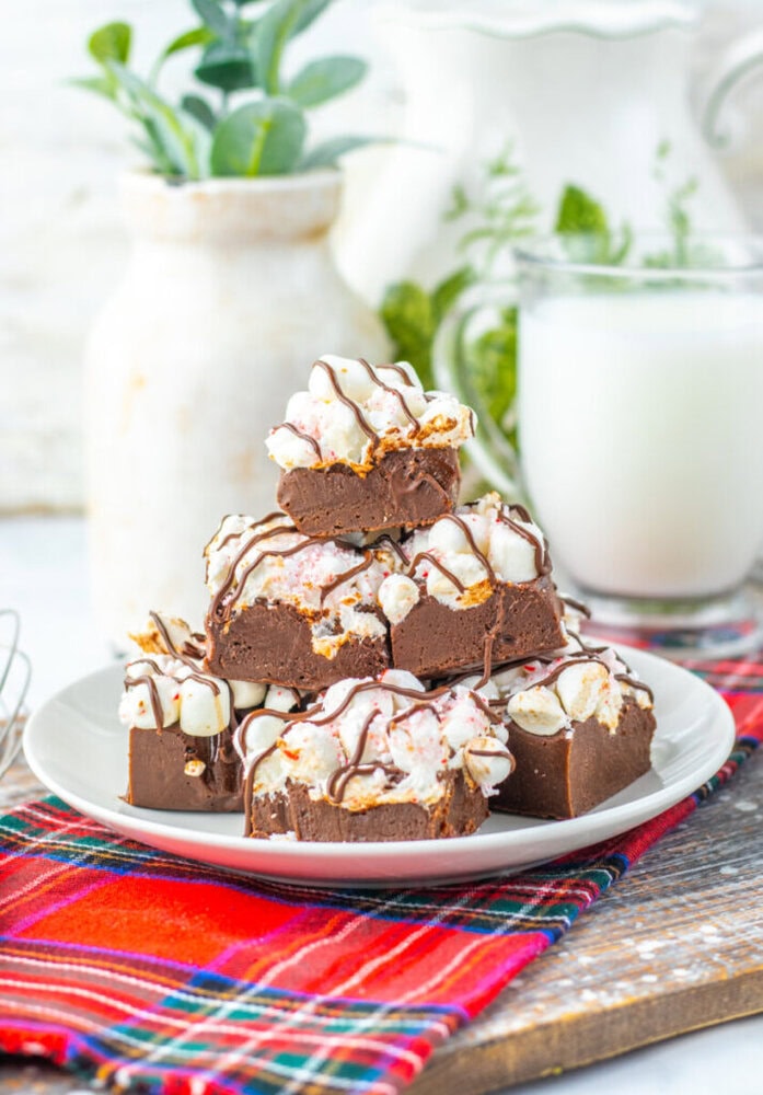 A plate stacked with chocolate fudge pieces topped with marshmallows and a white drizzle, set on a red plaid cloth, with a glass of milk and vase in the background.