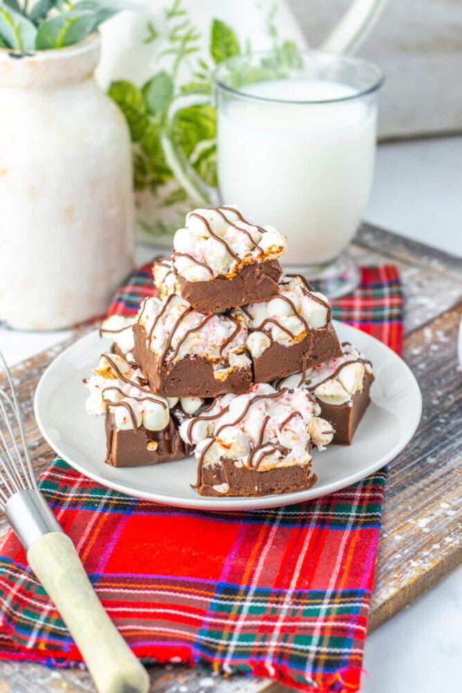 A plate with stacked chocolate bars topped with white and dark chocolate drizzle, set on a red plaid cloth, with a glass of milk and a whisk nearby.