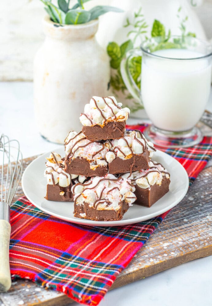 A plate of chocolate fudge squares with white and pink drizzle sits on a red plaid cloth, next to a glass of milk, a whisk, and a white vase with greenery.