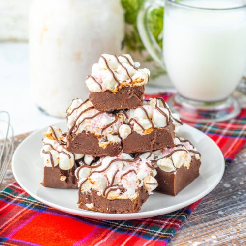 A plate of chocolate fudge topped with marshmallows and chocolate drizzle, set on a plaid cloth next to a glass of milk, a whisk, and a bowl of peppermint candies.