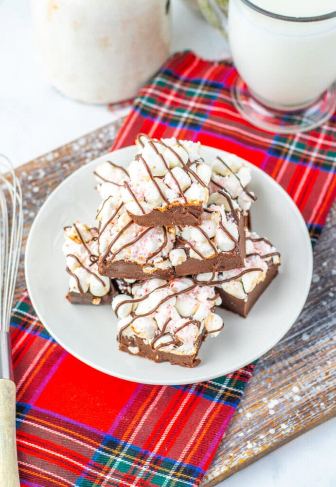 A plate of chocolate peppermint bark squares topped with white drizzle and crushed candy canes, set on a red plaid cloth next to a glass of milk and a whisk.