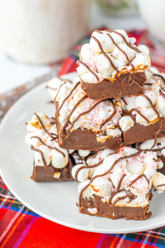 A plate of chocolate fudge squares topped with mini marshmallows and drizzled with chocolate sits on a white plate over a red plaid cloth.