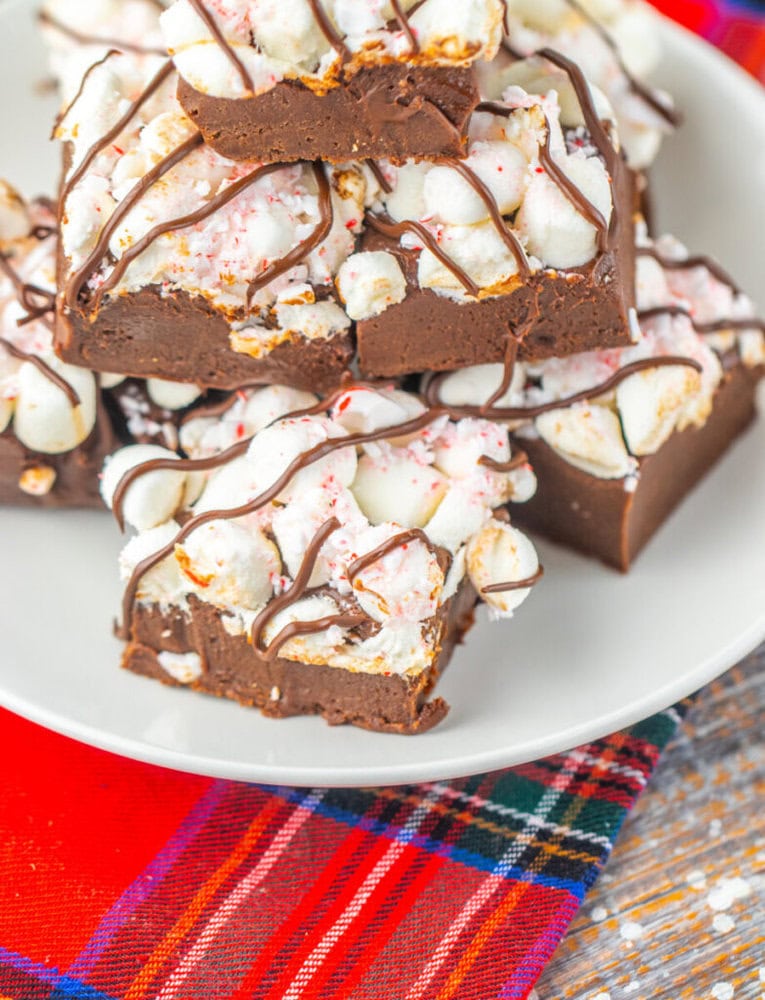 A plate of chocolate fudge squares topped with marshmallows and drizzled with chocolate, set on a red and plaid tablecloth.