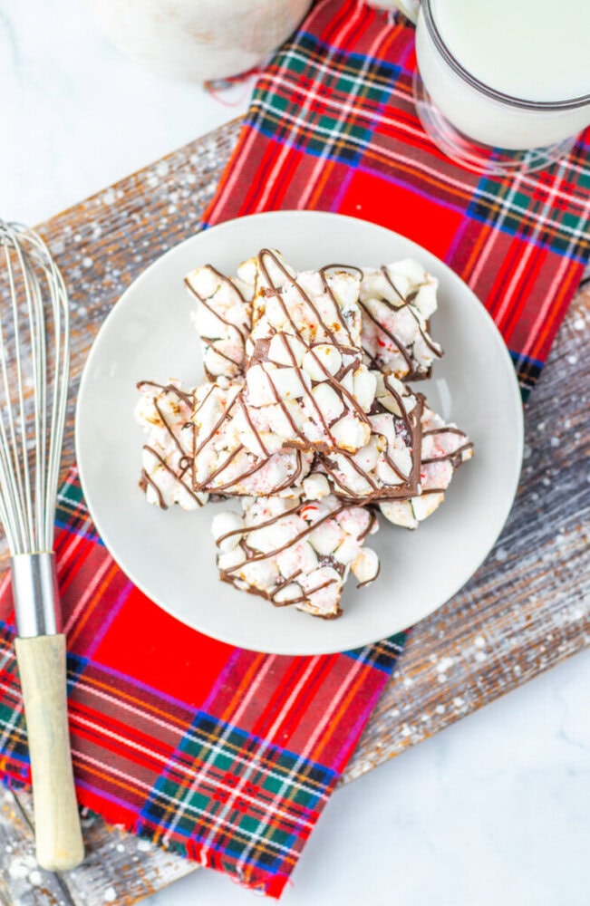 A plate of popcorn drizzled with chocolate sits on a wooden board with a red plaid cloth, next to a whisk and a glass of milk.