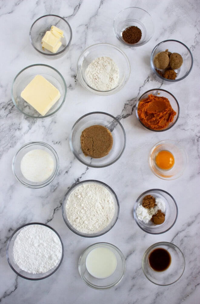 Assorted baking ingredients including flour, sugar, brown sugar, butter, egg yolk, pumpkin puree, spices, cocoa, milk, vanilla, and baking powder arranged in glass bowls on a marble surface.