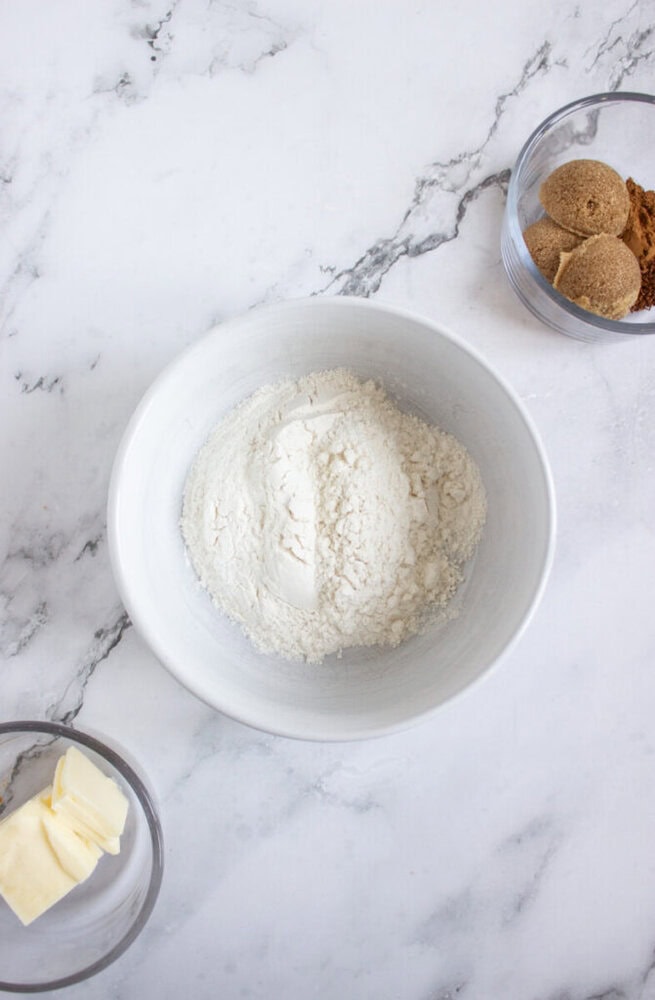 A white bowl with flour, a small glass bowl with sliced butter, and another bowl with brown sugar on a marble countertop.