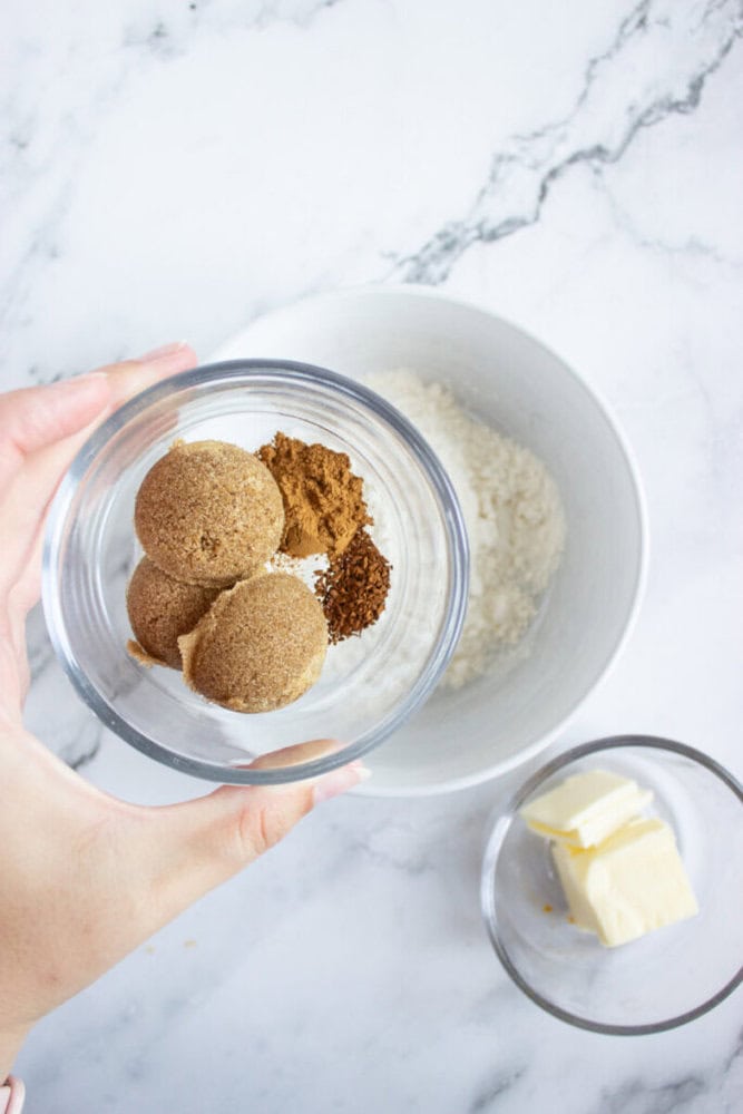 A hand holds a small bowl containing brown sugar and spices over a larger bowl of flour; a separate bowl with butter is nearby on a marble countertop.