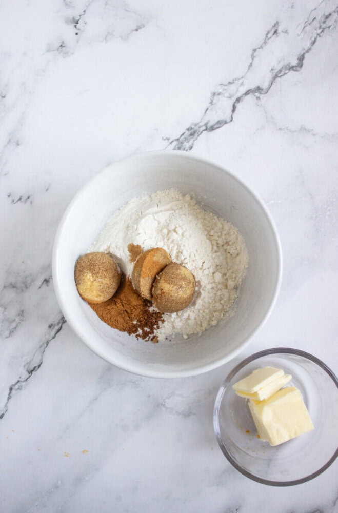 A white bowl contains flour, brown sugar, and cinnamon. Next to it, a small glass bowl holds several pieces of sliced butter on a marble surface.