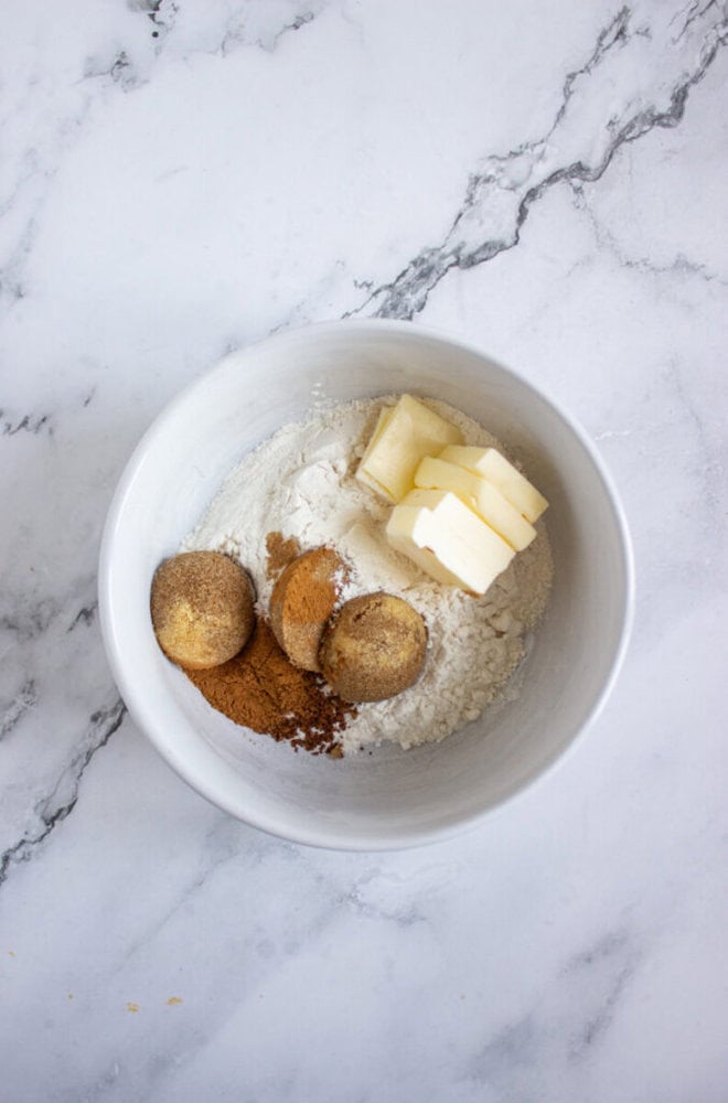 A white bowl on a marble surface contains flour, sliced butter, ground cinnamon, and brown sugar.