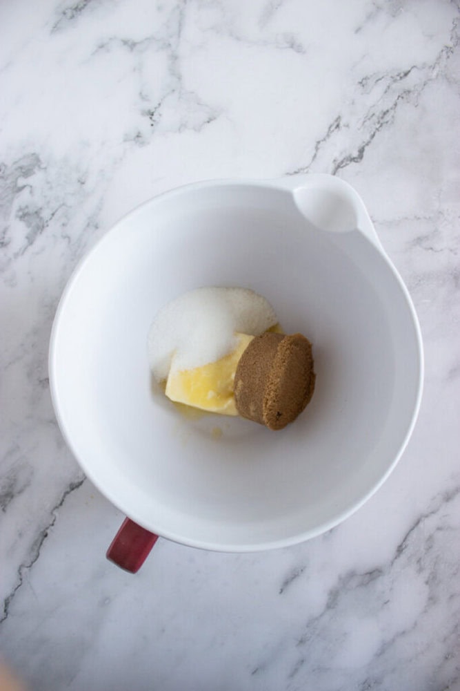 A white mixing bowl with a red handle containing butter, granulated sugar, and brown sugar, placed on a marble countertop.