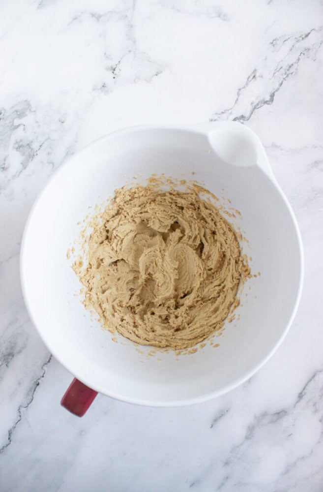 A white mixing bowl with light brown cookie dough batter sits on a marble countertop.