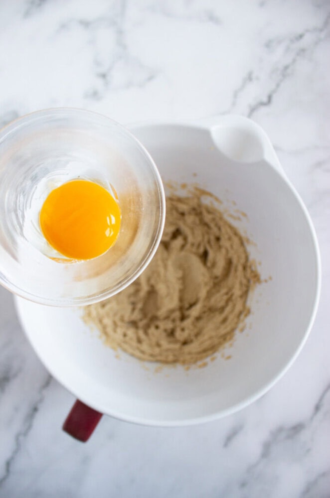 A glass cup holding a raw egg yolk is positioned above a white mixing bowl containing a creamed mixture on a marble countertop.