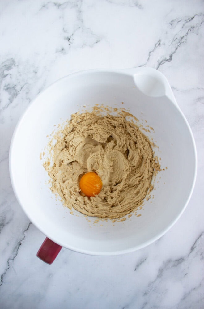 A white mixing bowl with creamed brown sugar and butter, and a cracked egg yolk in the center, sits on a marble countertop.