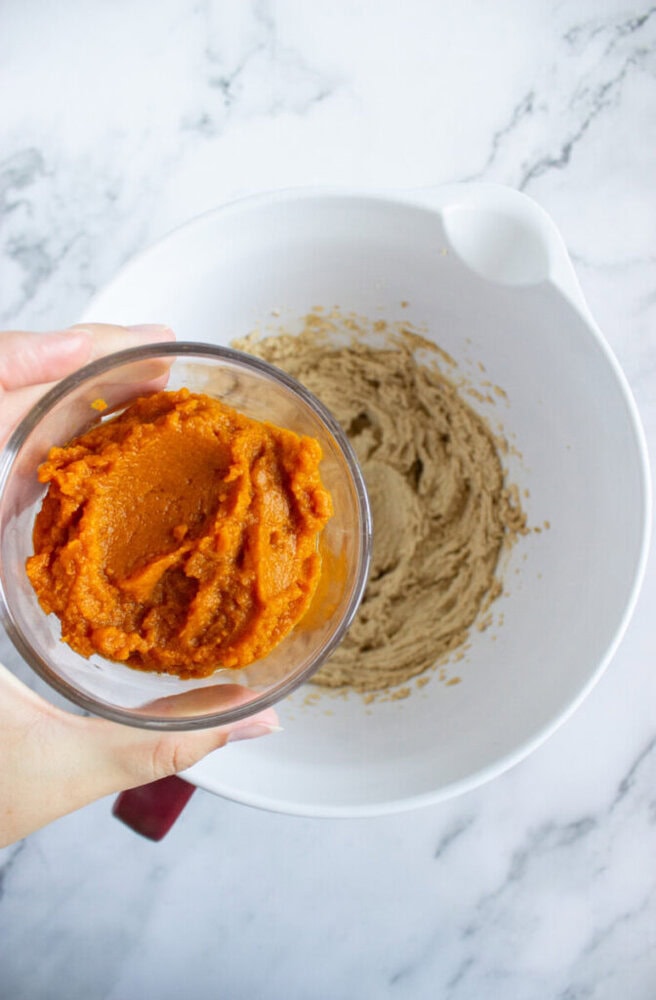 A hand holds a bowl of pumpkin puree above a mixing bowl containing creamed sugar and butter on a marble countertop.