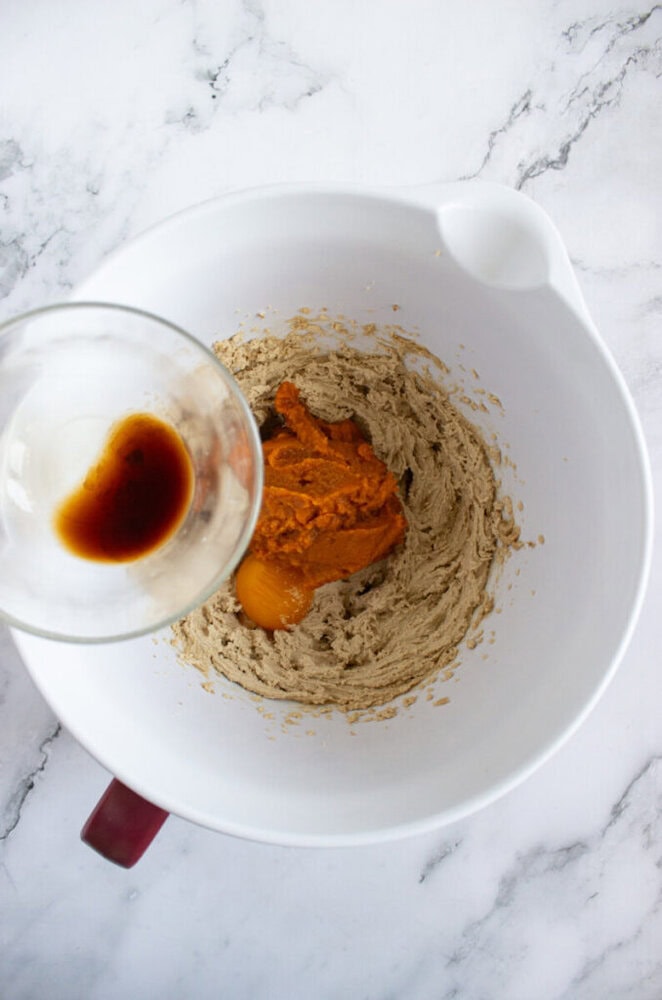 A white mixing bowl contains creamed sugar and butter, pumpkin puree, and an egg. A glass cup with vanilla extract is being poured in. The bowl is on a marble countertop.