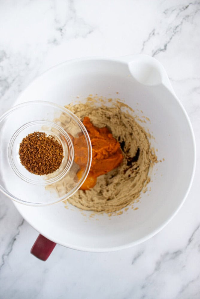 A glass bowl with instant coffee granules is held above a white mixing bowl containing pumpkin puree and creamed mixture on a marble countertop.