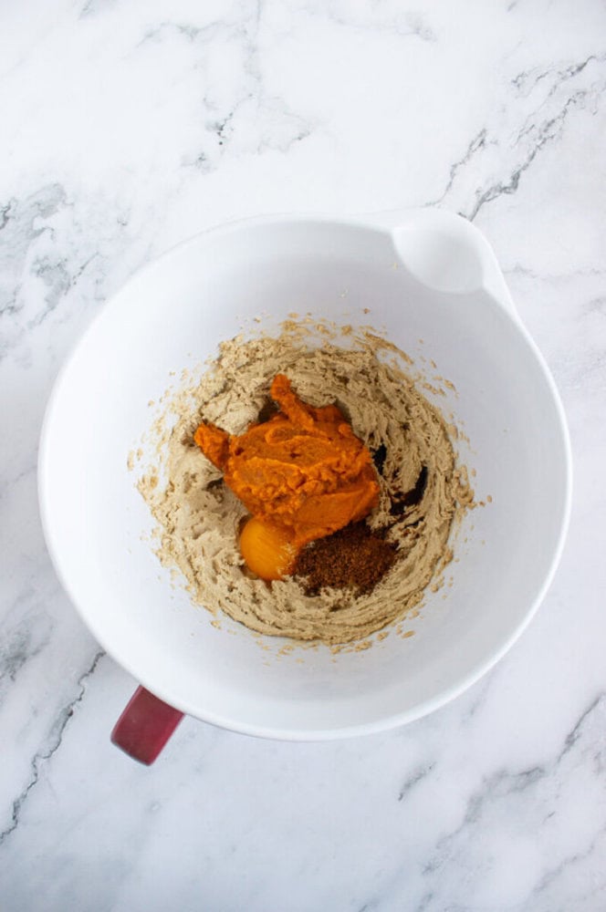 A white mixing bowl with brown sugar mixture, pumpkin puree, an egg, and spices on a marble countertop.