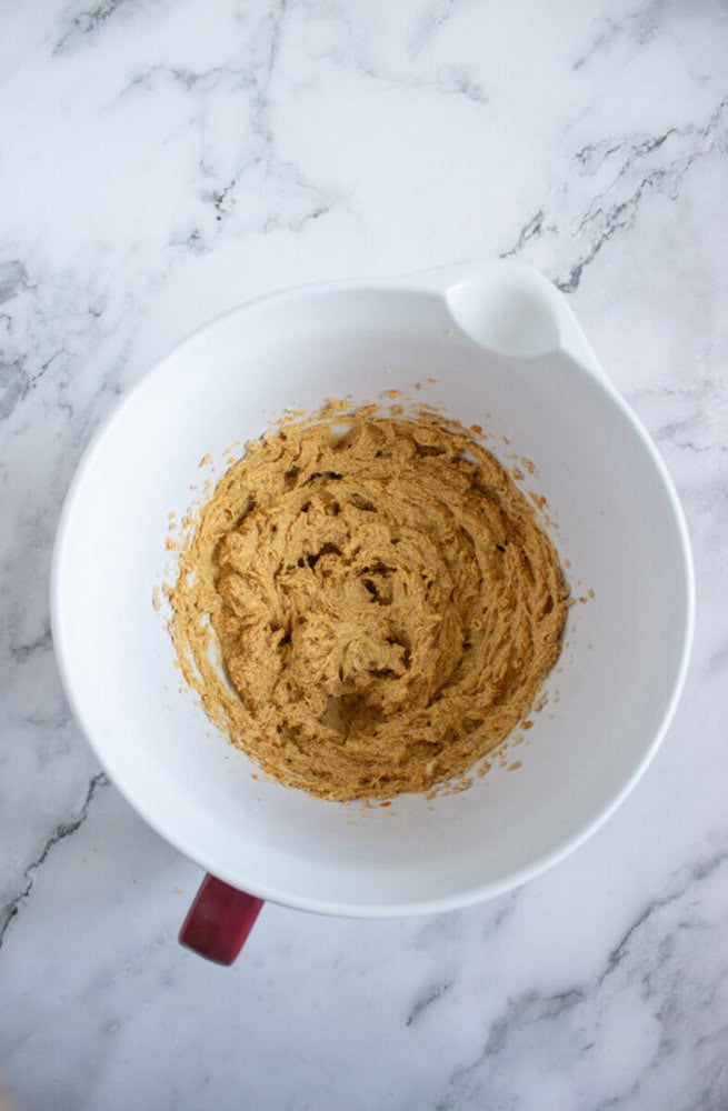 A white mixing bowl with a handle contains light brown cookie dough, sitting on a white marble countertop.