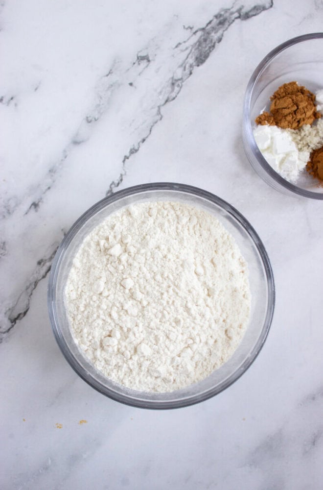A glass bowl filled with flour sits on a marble surface next to a smaller bowl containing brown and white powders.