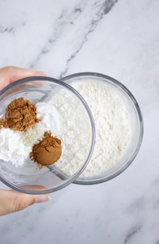 A hand holds a bowl containing cocoa powder, baking powder, and flour above another bowl filled with flour on a marble surface.