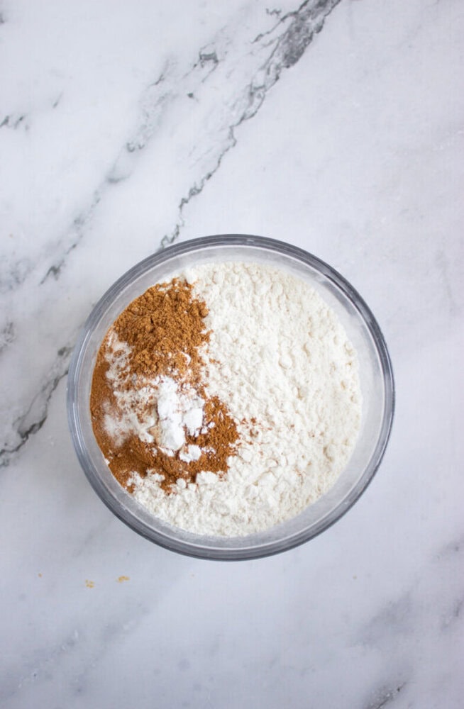 A glass bowl on a marble surface contains a mix of flour, ground cinnamon, and baking soda, with the ingredients separated but slightly overlapping.