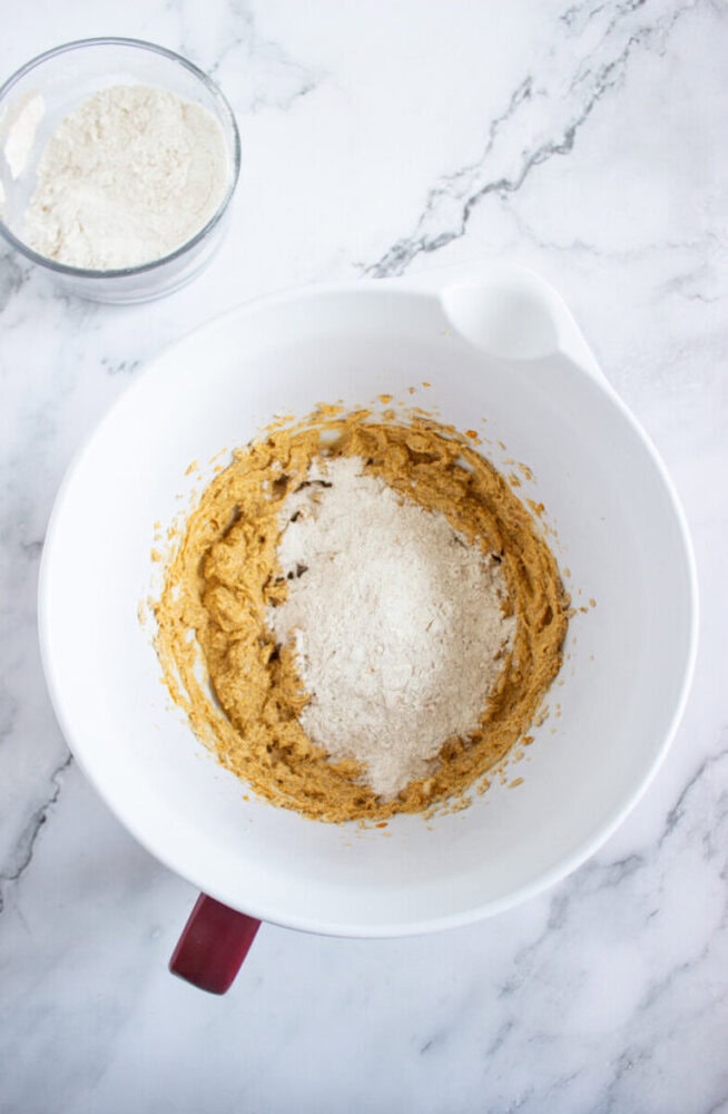 A white mixing bowl with a partially mixed batter and flour on top sits on a marble surface; a small bowl of flour is nearby.