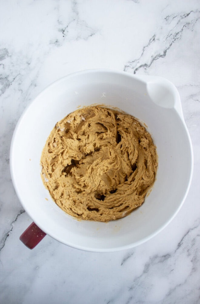 A white mixing bowl containing creamed brown cookie dough sits on a marble countertop.