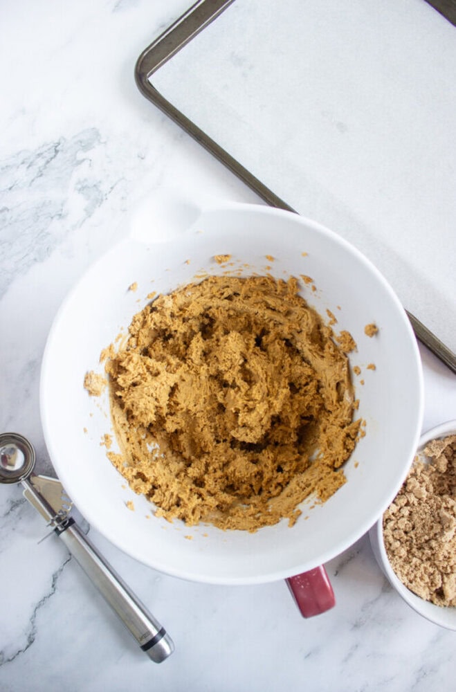 A mixing bowl with cookie dough, a cookie scoop, a bowl of brown sugar, and a baking sheet lined with parchment paper on a marble countertop.