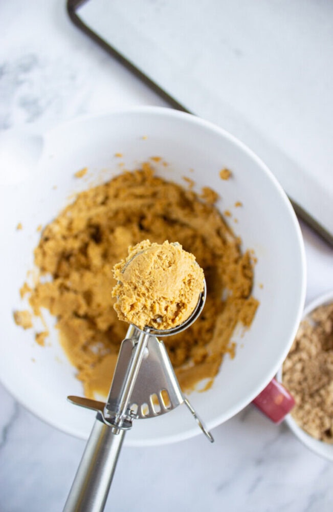 A metal cookie scoop holds a portion of cookie dough above a white mixing bowl filled with more dough. A baking sheet and a bowl of brown sugar are nearby.