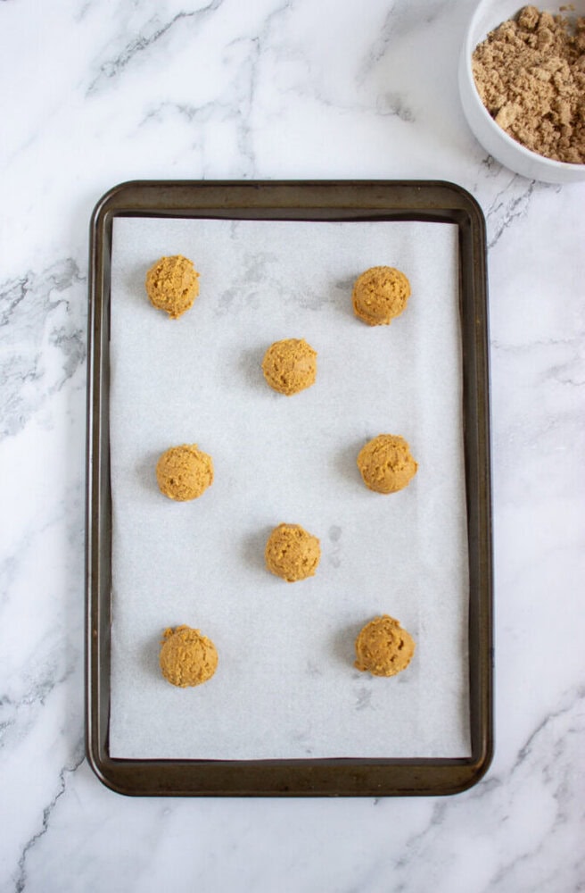 A baking tray lined with parchment paper holds nine evenly spaced scoops of cookie dough; a bowl of brown sugar sits nearby on a marble surface.