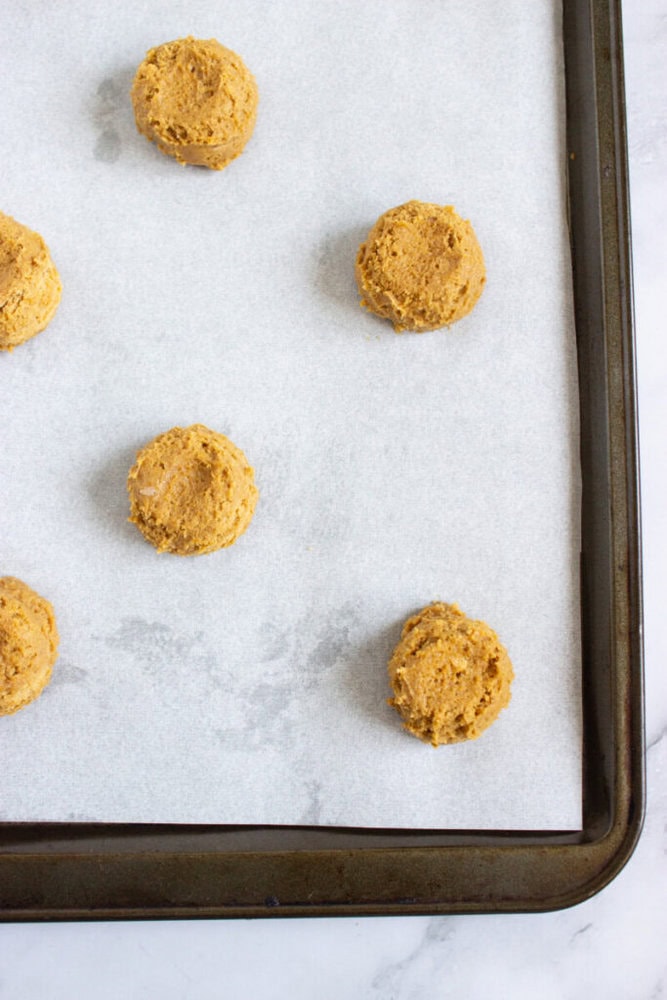 Six unbaked cookie dough balls are arranged on a parchment-lined baking sheet.