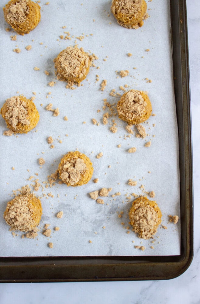 A baking sheet lined with parchment paper holds spaced, unbaked cookie dough balls topped with crumbly streusel.