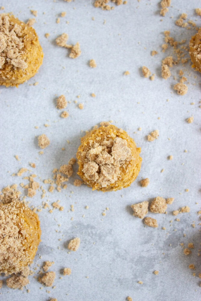 Scoops of cookie dough topped with brown sugar are arranged on a parchment-lined baking sheet, with some loose brown sugar scattered around.