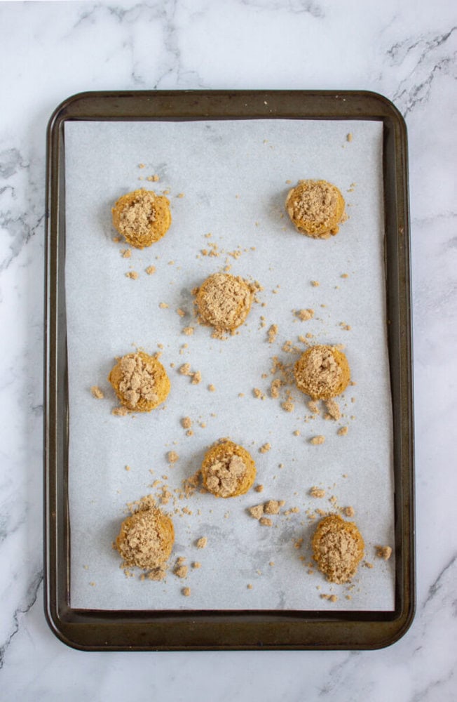 A baking sheet lined with parchment paper holds eight unbaked dough balls sprinkled with crumb topping, on a marble surface.