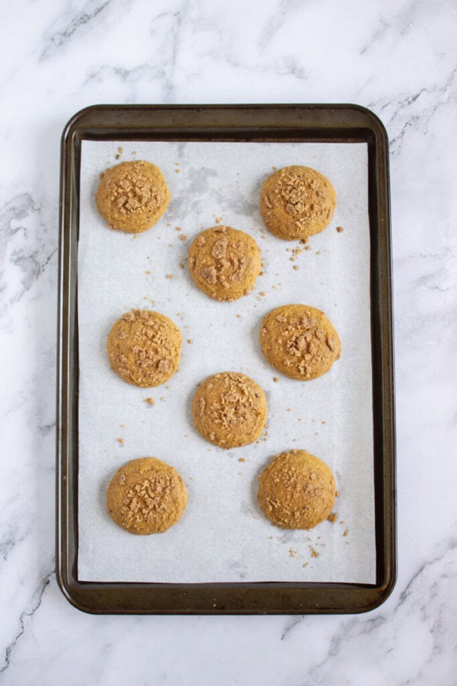 A baking tray lined with parchment paper holds eight round cookies, evenly spaced, with a crumbly topping. The tray is on a white marble surface.