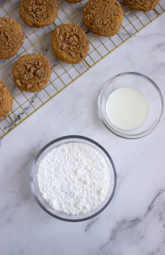 A cooling rack with crumb-topped muffins next to bowls of powdered sugar and milk on a marble surface.