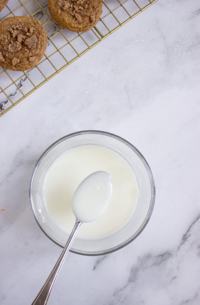 A bowl of white glaze with a spoon rests on a marble surface; muffins are cooling on a wire rack in the background.