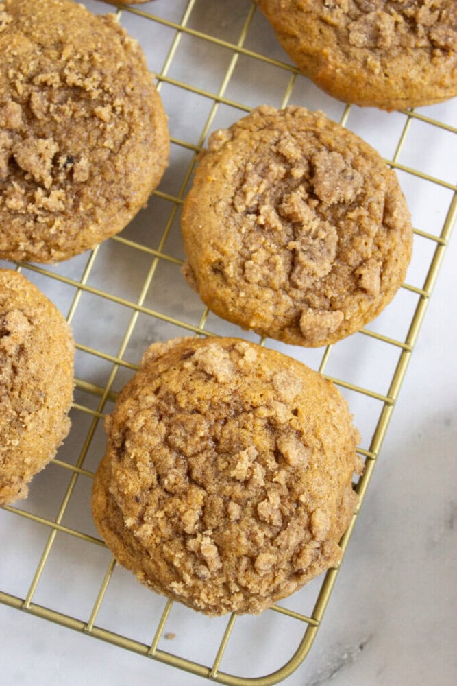 Pumpkin muffins with a crumb topping are cooling on a wire rack placed on a white surface.