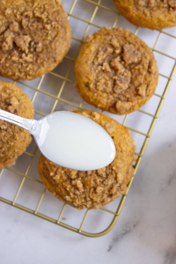 A spoonful of white glaze is held over pumpkin streusel muffins on a wire cooling rack.