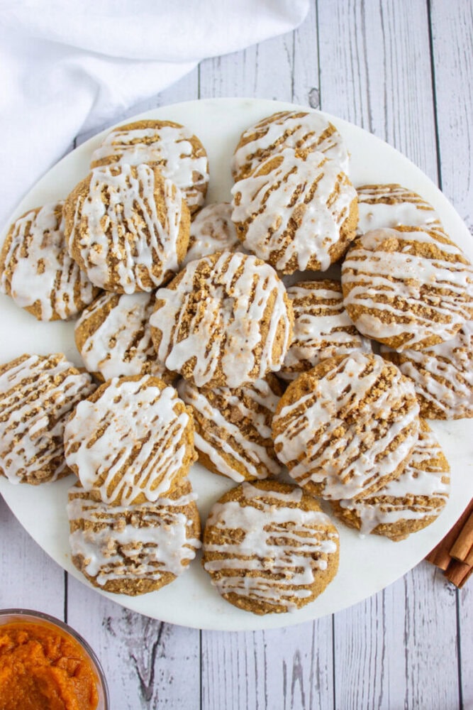 A plate of round cookies with white icing drizzled on top, arranged on a white marble surface, with a small bowl of orange spread nearby.