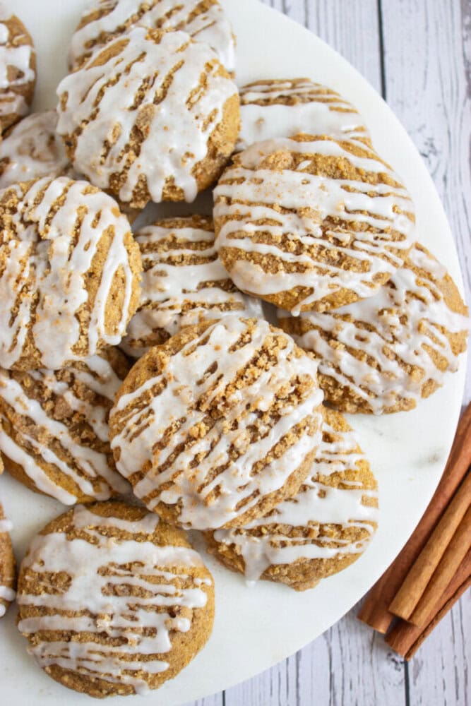 A plate of round, iced cookies with a drizzle of white glaze sits on a white surface next to a few cinnamon sticks.