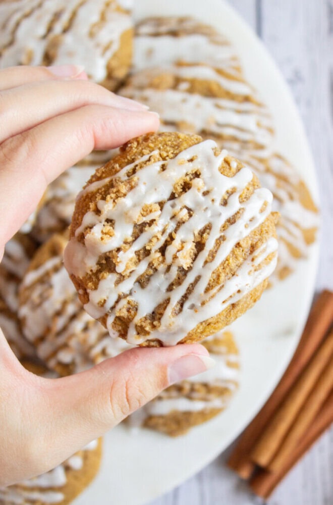 A hand holds a round cookie topped with white icing drizzle; more cookies and cinnamon sticks are on a plate in the background.