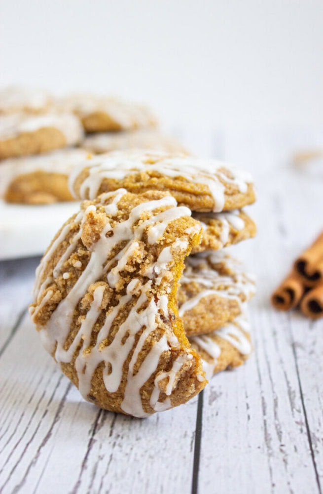 A stack of glazed cookies with one cookie in front showing a bite taken out, set on a white wooden surface with cinnamon sticks in the background.