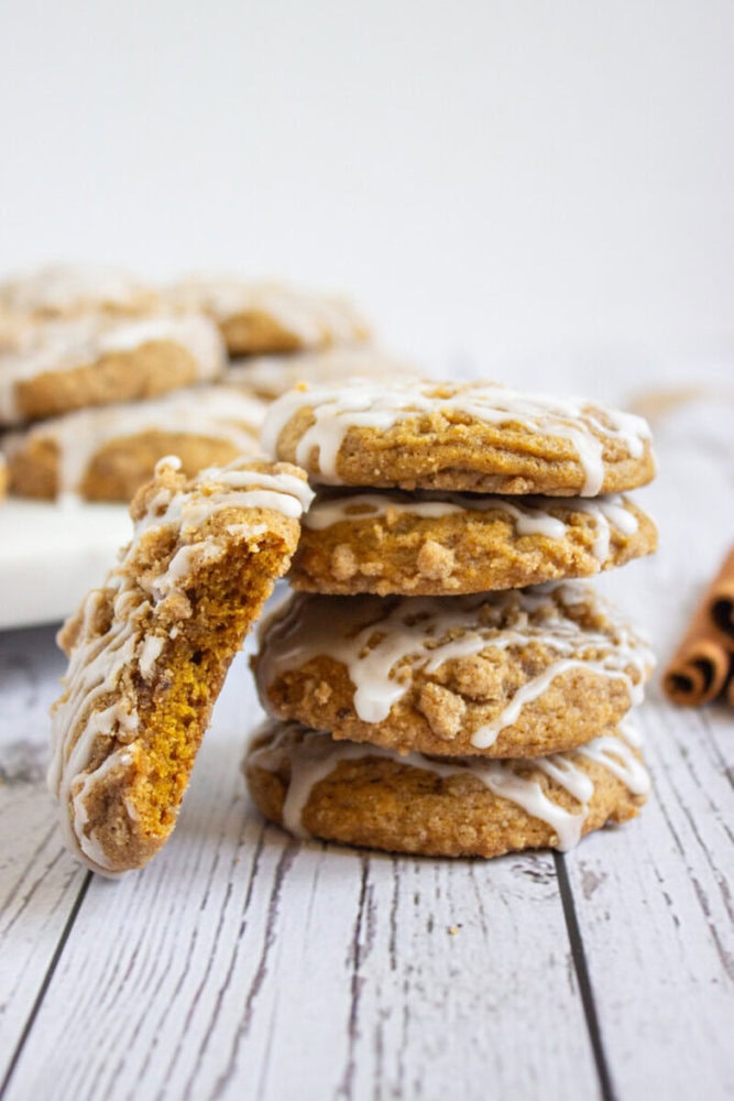 A stack of four iced cookies sits on a white wooden surface, with one cookie partially eaten and more cookies in the background.