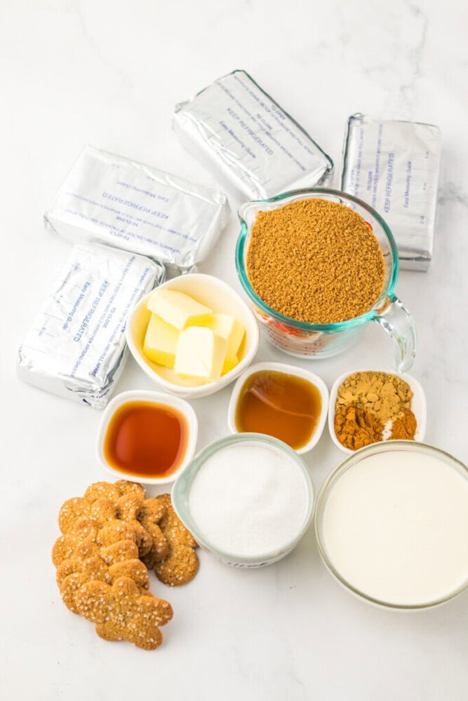 Ingredients for a cheesecake recipe arranged on a white surface, including cream cheese, brown sugar, butter, vanilla extract, spices, granulated sugar, cream, and ginger snap cookies.